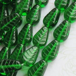 May include: Close-up of several green leaf-shaped glass beads. Each bead has a detailed leaf vein pattern. The beads are a vibrant emerald green color and appear to be strung together. These beads are suitable for jewelry making and crafting.