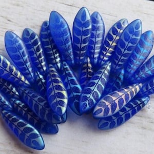 May include: A collection of blue, leaf-shaped beads with a silver leaf pattern. The beads are arranged in a cluster, showcasing their iridescent quality. The beads are likely for jewelry making or craft projects.
