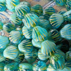 May include: A collection of translucent turquoise glass beads shaped like seashells. Each bead features a series of raised, gold-colored lines radiating from the base to the edge, creating a textured, fan-like pattern across the surface.
