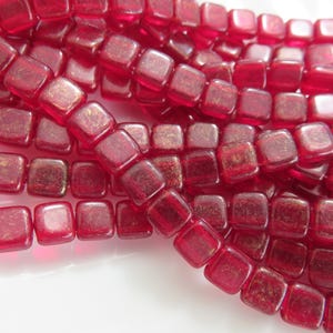 May include: Close-up of strands of red square beads. The beads have a translucent quality with a hint of gold flecks. The beads are strung together, creating a textured and visually appealing pattern.