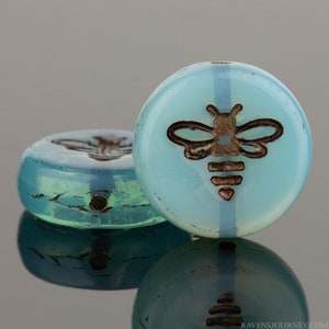 May include: Two round glass beads with a light blue and white opalescent finish. The beads feature a brown bee design.
