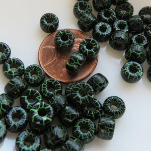 May include: A collection of black beads with a green crackle pattern. The beads are various shapes, including round and cylindrical, and are scattered on a white surface. A penny is included for size reference.