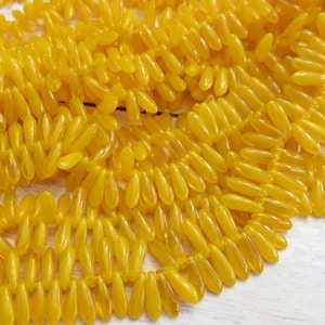 May include: Close-up of a collection of yellow, teardrop-shaped beads. The beads are strung together, creating a textured surface. The beads have a glossy finish and are arranged in a pattern, suggesting they are for jewelry making.