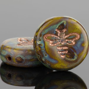 May include: Two round ceramic beads with a copper-colored bee design. The beads have a mottled green and brown glaze.
