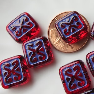 May include: Close-up of several red square glass beads with a blue and white decorative design. The beads are next to a penny for size comparison. The beads have a textured surface and are on a white background.
