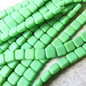 May include: A strand of square-shaped, matte green glass beads. The beads are strung on a clear thread and are arranged in a staggered pattern.