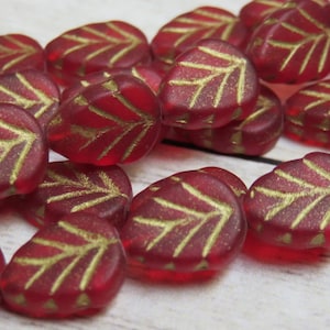 May include: A close-up of red glass beads with gold leaf accents in the shape of leaves. The beads are scattered on a light wood surface.