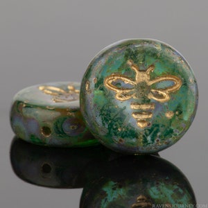 May include: Two green glass beads with a gold bee design. The beads have a mottled, iridescent finish.