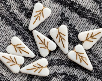 Op de afbeelding: Witte hartvormige kralen met goudkleurige bladeren. De kralen zijn gerangschikt op een gestructureerd grijs en wit oppervlak. Deze decoratieve kralen zijn ideaal voor het maken van sieraden en knutselprojecten.
