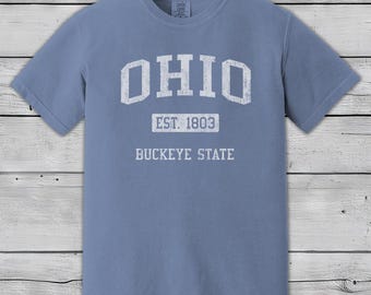 Könnte beinhalten: Ein blaues T-Shirt mit dem Wort "OHIO" in großen weißen Buchstaben, zusammen mit "EST. 1803" und "BUCKEYE STATE" in weißem Text. Das Shirt hat kurze Ärmel und einen Rundhalsausschnitt. Der Markenname "Jim Shorts" ist oben links aufgedruckt.