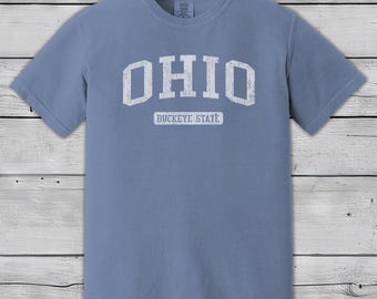 Könnte beinhalten: Staubblaues T-Shirt mit dem Wort "OHIO" in großen, weißen Buchstaben auf der Brust. Darunter steht "BUCKEYE STATE" in einem kleineren, rechteckigen weißen Feld. Das Shirt hat kurze Ärmel.
