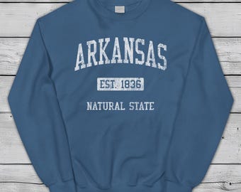 Può includere: Felpa blu con la scritta bianca "ARKANSAS". Sotto, "EST. 1836" e "NATURAL STATE" sempre bianche. Il marchio "Jim Shorts" è in alto a sinistra. Design vintage.