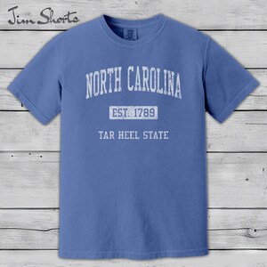 K&ouml;nnte beinhalten: Ein blaues T-Shirt mit dem wei&szlig;en Schriftzug "NORTH CAROLINA" oben. Darunter steht "EST. 1789" und "TAR HEEL STATE". Das Shirt hat kurze &Auml;rmel und einen Rundhalsausschnitt. Der Markenname "Jim Shorts" befindet sich oben links.