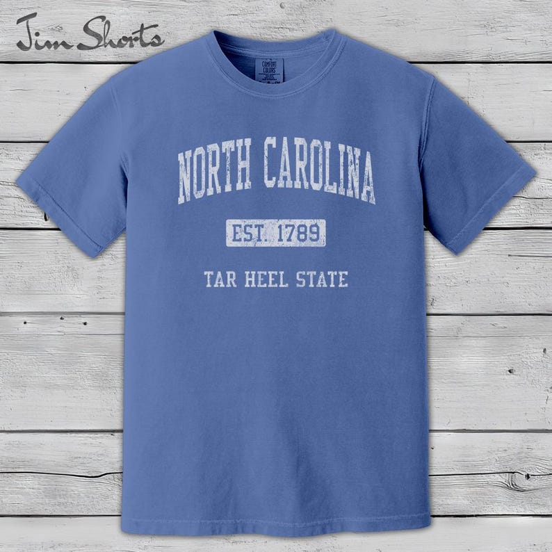 K&ouml;nnte beinhalten: Ein blaues T-Shirt mit dem wei&szlig;en Schriftzug "NORTH CAROLINA" oben. Darunter steht "EST. 1789" und "TAR HEEL STATE". Das Shirt hat kurze &Auml;rmel und einen Rundhalsausschnitt. Der Markenname "Jim Shorts" befindet sich oben links.