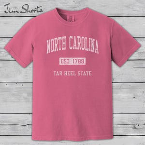 K&ouml;nnte beinhalten: Ein pinkfarbenes T-Shirt mit dem wei&szlig;en Schriftzug "NORTH CAROLINA" oben. Darunter steht zentriert "EST. 1789" und unten "TAR HEEL STATE". Das Shirt hat kurze &Auml;rmel und einen Rundhalsausschnitt.