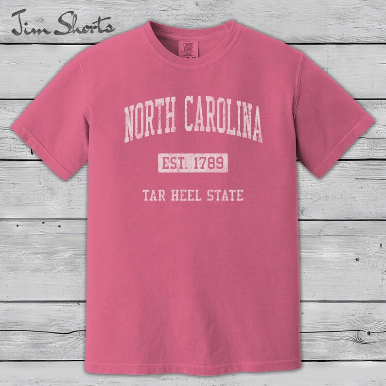 K&ouml;nnte beinhalten: Ein pinkfarbenes T-Shirt mit dem wei&szlig;en Schriftzug "NORTH CAROLINA" oben. Darunter steht zentriert "EST. 1789" und unten "TAR HEEL STATE". Das Shirt hat kurze &Auml;rmel und einen Rundhalsausschnitt.
