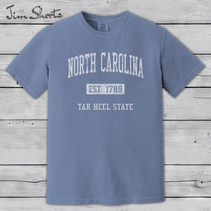 K&ouml;nnte beinhalten: Hellblaues T-Shirt mit wei&szlig;em Aufdruck "NORTH CAROLINA". Darunter steht "EST. 1789" und "TAR HEEL STATE". Das T-Shirt hat kurze &Auml;rmel und einen Rundhalsausschnitt.