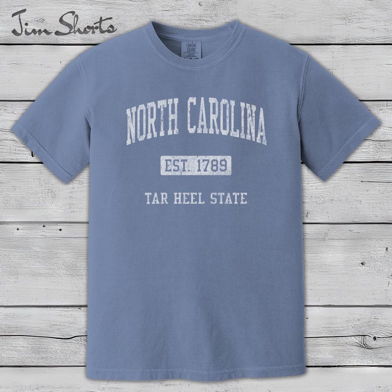 K&ouml;nnte beinhalten: Hellblaues T-Shirt mit wei&szlig;em Aufdruck "NORTH CAROLINA". Darunter steht "EST. 1789" und "TAR HEEL STATE". Das T-Shirt hat kurze &Auml;rmel und einen Rundhalsausschnitt.