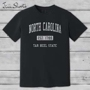 K&ouml;nnte beinhalten: Schwarzes T-Shirt mit wei&szlig;em Text: "NORTH CAROLINA", "EST. 1789" und "TAR HEEL STATE". Das T-Shirt hat einen Rundhalsausschnitt und kurze &Auml;rmel. Der Hintergrund ist Holz.