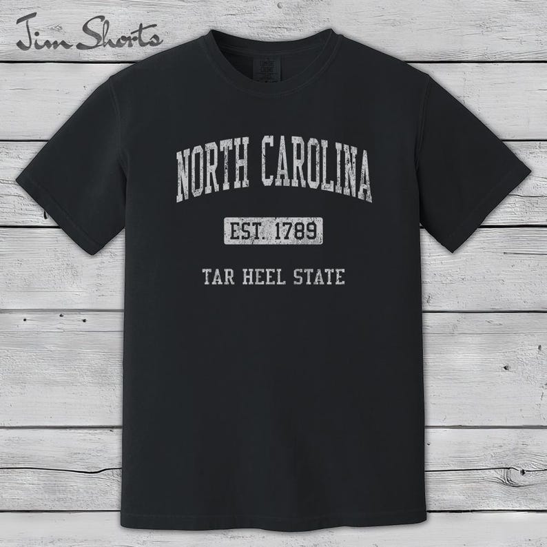 K&ouml;nnte beinhalten: Schwarzes T-Shirt mit wei&szlig;em Text: "NORTH CAROLINA", "EST. 1789" und "TAR HEEL STATE". Das T-Shirt hat einen Rundhalsausschnitt und kurze &Auml;rmel. Der Hintergrund ist Holz.
