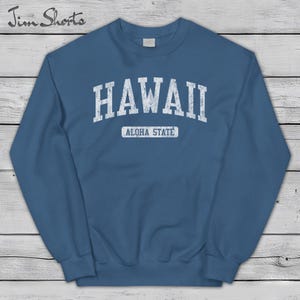 May include: A blue sweatshirt with the word "HAWAII" in distressed white lettering across the chest. Below the word "HAWAII" is the text "ALOHA STATE" in a smaller white font. The sweatshirt has a crew neck and long sleeves.