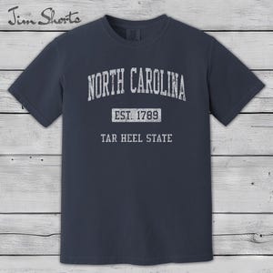 K&ouml;nnte beinhalten: Ein marineblaues T-Shirt mit dem wei&szlig;en Text "NORTH CAROLINA" oben in einem Bogen. Darunter steht "EST. 1789" und "TAR HEEL STATE". Das T-Shirt hat einen Rundhalsausschnitt und kurze &Auml;rmel.