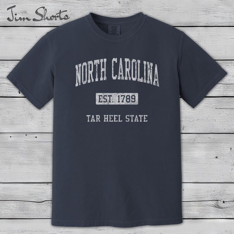 K&ouml;nnte beinhalten: Ein marineblaues T-Shirt mit dem wei&szlig;en Text "NORTH CAROLINA" oben in einem Bogen. Darunter steht "EST. 1789" und "TAR HEEL STATE". Das T-Shirt hat einen Rundhalsausschnitt und kurze &Auml;rmel.