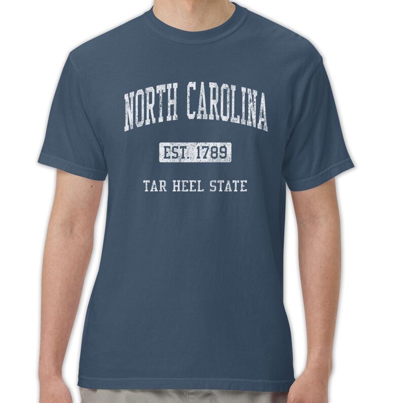 K&ouml;nnte beinhalten: Marineblaues T-Shirt mit dem Text "NORTH CAROLINA" in verblichener wei&szlig;er Schrift. Darunter steht "EST. 1789" zentriert, gefolgt von "TAR HEEL STATE". Das T-Shirt hat kurze &Auml;rmel und eine klassische Passform.