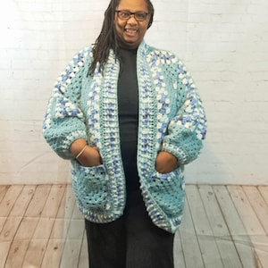 May include: A light blue crocheted cardigan with a white and blue geometric pattern. The cardigan has a shawl collar and two pockets.