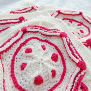 Op de afbeelding: Witte gehaakte vierkanten met roze bies en roze bloemaccenten. De vierkanten zijn op elkaar gelegd.