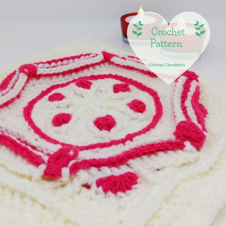 Op de afbeelding: Een wit en roze gehaakt vierkant met een cirkelvormig ontwerp in het midden. Het vierkant heeft een rand van witte en roze strepen. Het middelste ontwerp heeft een cirkel met roze stippen en een roze hart in het midden. De tekst "Crochet Pattern" staat in een hartvorm boven het vierkant.