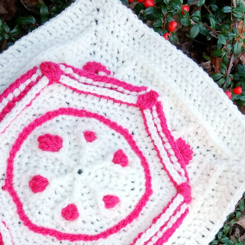 Op de afbeelding: Wit gehaakt vierkant met een roze en wit geometrisch ontwerp in het midden. Het ontwerp heeft een cirkel met zes roze pompoenen rond de rand.