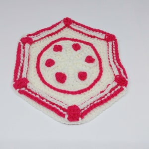 Op de afbeelding: Een wit gehaakt hexagon met een roze gehaakt ontwerp in het midden. Het ontwerp heeft een cirkel met vijf roze cirkels erin.