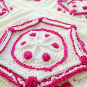 Op de afbeelding: Close-up van een gehaakt deken met een witte achtergrond en roze accenten. Het deken heeft een herhalend patroon van geometrische vormen met een centrale cirkel.