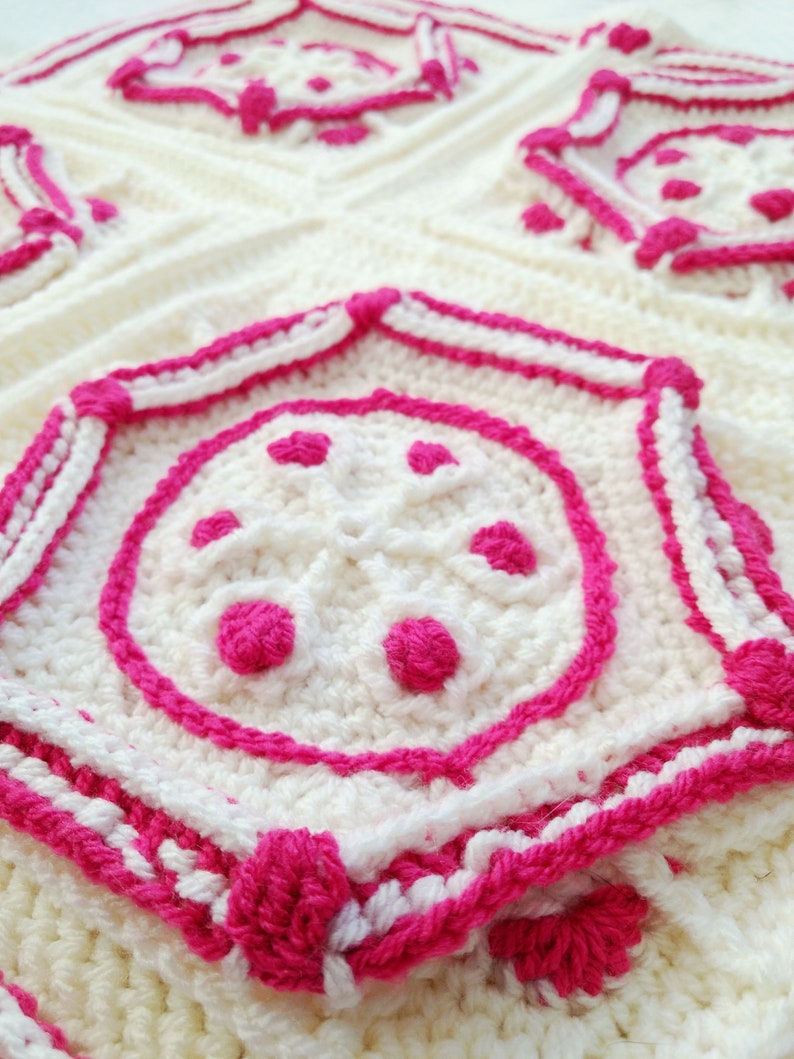Op de afbeelding: Close-up van een gehaakt deken met een witte achtergrond en roze accenten. Het deken heeft een herhalend patroon van geometrische vormen met een centrale cirkel.
