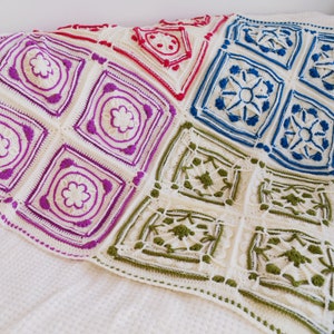 Op de afbeelding: Een wit gehaakt deken met kleurrijke vierkanten. De vierkanten zijn gemaakt in verschillende patronen en kleuren, waaronder roze, blauw, groen en rood.