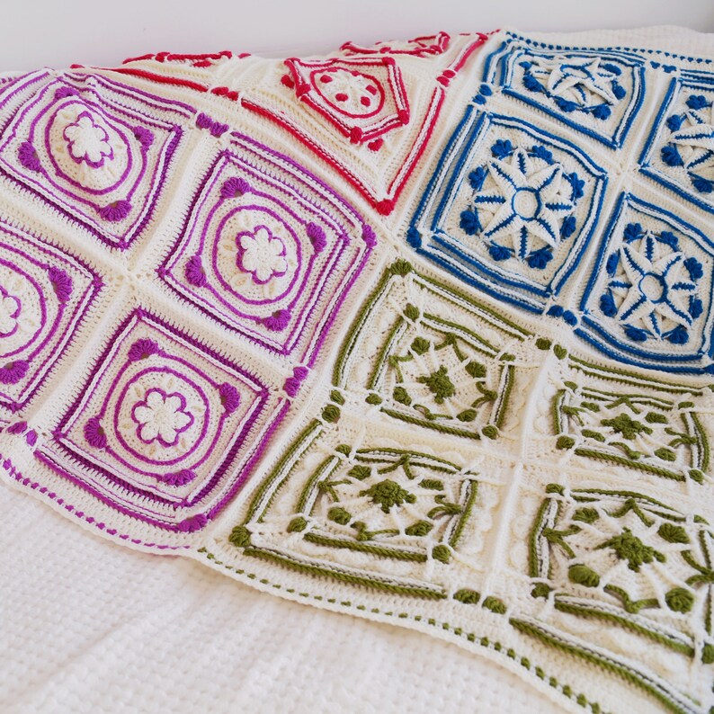 Op de afbeelding: Een wit gehaakt deken met kleurrijke vierkanten. De vierkanten zijn gemaakt in verschillende patronen en kleuren, waaronder roze, blauw, groen en rood.