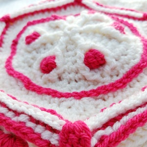 Op de afbeelding: Close-up van een wit gehaakt vierkant met een roze rand. Het midden van het vierkant heeft een verhoogd, bloemvormig ontwerp met drie roze cirkels.