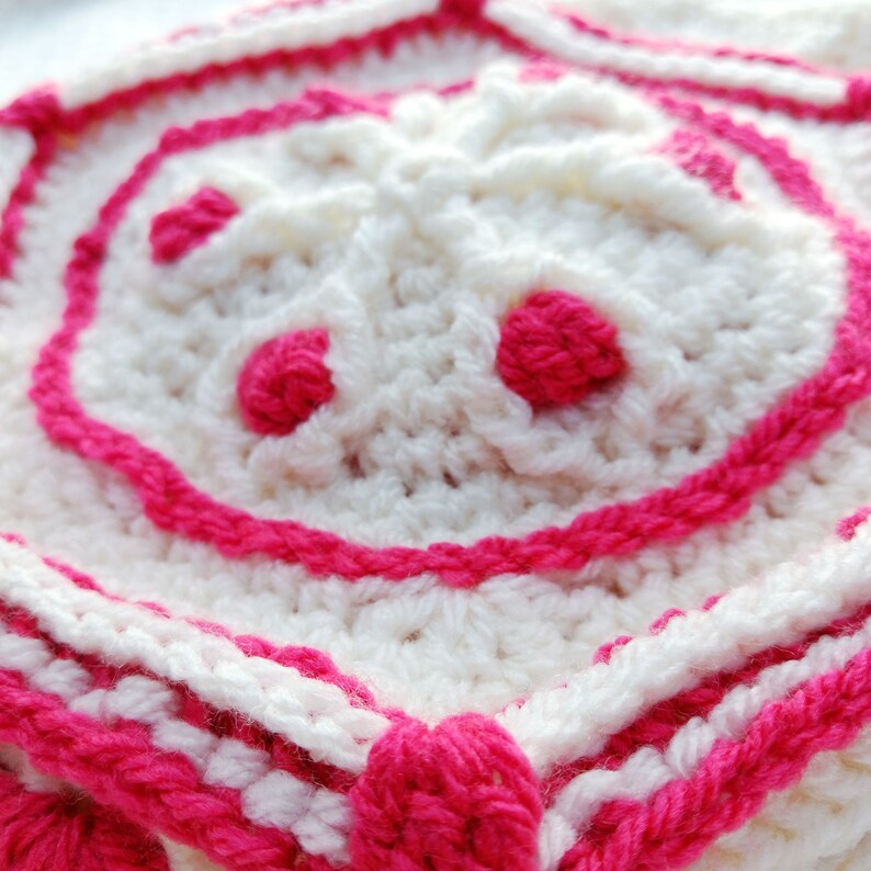 Op de afbeelding: Close-up van een wit gehaakt vierkant met een roze rand. Het midden van het vierkant heeft een verhoogd, bloemvormig ontwerp met drie roze cirkels.