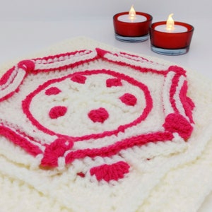 Op de afbeelding: Een wit gehaakt vierkant met een roze gehaakt ontwerp in het midden. Het ontwerp heeft een cirkel met roze stippen en een roze rand.