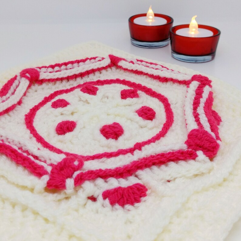 Op de afbeelding: Een wit gehaakt vierkant met een roze gehaakt ontwerp in het midden. Het ontwerp heeft een cirkel met roze stippen en een roze rand.