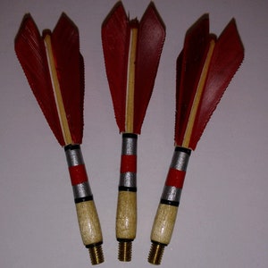 May include: Three red and white feathered darts with silver and red bands on the wooden shafts. The darts have brass tips.