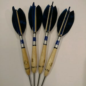 Puede incluir: Cuatro dardos de plumas azules y negras con ejes de madera y bandas plateadas y azules. Los ejes están etiquetados como "Belgian Darts".