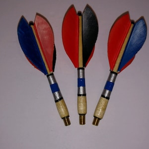 Puede incluir: Tres dardos con plumas rojas, azules y negras. Los dardos tienen ejes de madera y puntas de latón.