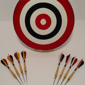 Puede incluir: Un tablero de dardos rojo, blanco y negro con un centro y ocho dardos con plumas de colores.