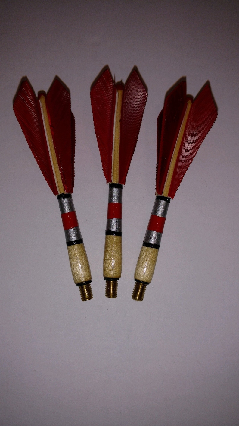 May include: Three red and white feathered darts with silver and red bands on the wooden shafts. The darts have brass tips.