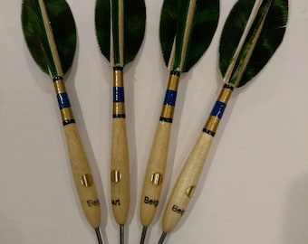 May include: Four green and white feathered darts with gold and blue accents and wooden shafts. The darts have the word "Beige" written on them.