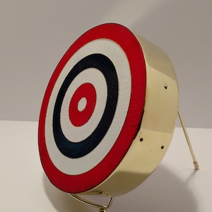 Pode incluir: Um alvo de metal dourado com círculos concêntricos vermelhos, brancos e pretos. O alvo está montado em um suporte de metal dourado com uma base redonda.
