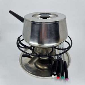 May include: A silver fondue pot with a black handle and a black lid. The pot is sitting on a silver base with a black metal ring around it. There are six black fondue forks with colored tips.