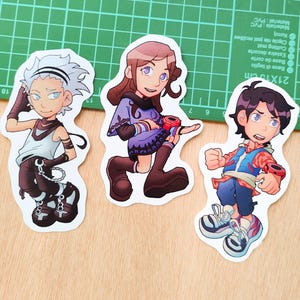 Puede incluir: Tres pegatinas de personajes de estilo anime. Los personajes están representados en poses dinámicas, con ropa y accesorios detallados. Las pegatinas están sobre una superficie de madera, con una esterilla de corte verde en el fondo. Los personajes tienen diversos peinados y vestimentas.